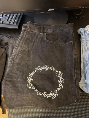 Svarta jeans med vit textcirkel - Unika svarta jeans med vita cirklar av texten 'Eightyfive' tryckt på flera ställen. Rak passform och klassisk femficksmodell. Jeansen har bälteshällor och dragkedjegylf. Materialet är denim i bomull och designen sticker ut med sin grafiska stil.