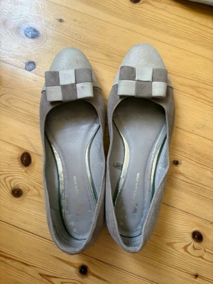 Beige ballerinaskor med rutigt band - Säljer ett par beige ballerinaskor i mockaimitation med rund tå och dekorativt rutigt band framtill i beige och ljusgrått. Skorna har en klassisk och stilren design som passar till många olika outfits. Säljer då de tyvärr är för stora💕🌺