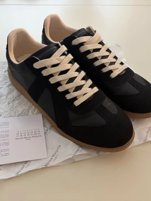 Maison Margiela Gats svarta sneakers - Svarta gats sneakers från Maison Margiela med beigea skosnören och gummisula. Skorna har en mix av mocka och läderdetaljer, klassisk låg silhuett och diskret design som passar till det mesta. Hör gärna av er vid bud eller frågor:)