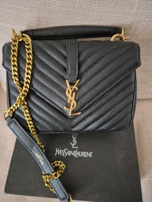 Svart Axelväska  - Snygg svart handväska från Ysl i quiltat läder med guldfärgad kedja och ikonisk YSL-logga framtill. Väskan har ett top handtag och axelrem i kedja, perfekt för en trendig look. Klassisk rektangulär form och elegant design.
