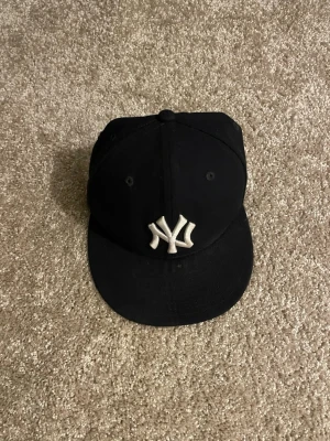 Svart New York Yankees keps - Snygg svart keps med klassiskt New York Yankees-broderi i vitt framtill. Kepsen har böjd skärm och är tillverkad i ett slitstarkt tyg. Perfekt för dig som vill ha en stilren och sportig look.