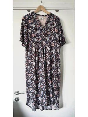 Noa Noa - Blommig midiklänning med v-ringning - storlek 38 - Härlig klänning från Noa Noa i storlek 38 med ett detaljerat blommönster i olika nyanser av blått, rosa och brunt mot en mörkblå bakgrund. Tidlös modell med korta ärmar och en klassisk krage med V-ringning. Klänningen knäpps med knappar framtill och har en rundad nederkant med slitsar i sidorna. Luftigt finvävt tyg i 100% viskos, så bra naturmaterial som andas. Bekväm och vacker klänning som är lika enkel att styla upp för fest, som ned för jobb / vardag. Nypris 1249:-. Mycket gott skick.