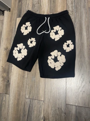 Svarta shorts med blommigt mönster - Svarta shorts med vita blommiga tryck och vit snörning i midjan. Perfekta för sommaren och har en avslappnad passform. Snyggt och trendigt plagg för en chill stil.