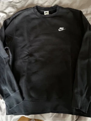 Svart sweatshirt från Nike - Klassisk svart sweatshirt från Nike med vit broderad logga på bröstet. Tröjan har rund halsringning, långa ärmar och ribbade muddar vid ärmslut och nederkant. Perfekt för en avslappnad och sportig stil.