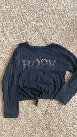 Svart långärmad topp med knyt - Cool svart långärmad topp från Firefly med texten HOPE i guldiga prickar framtill. Toppen har ett knyt i nederkanten för en trendig look och är gjord i mjuk bomull. Perfekt för dig som vill ha en chill men snygg stil.