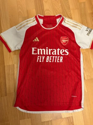Arsenal matchtröja röd/vit - Snygg Arsenal matchtröja från Adidas i rött och vitt med gulddetaljer på axlarna. Tröjan har klubbmärke och sponsortryck framtill samt ett mönster av tunna svarta blixtar. Tillverkad i lätt och ventilerande funktionsmaterial, perfekt för fotbollsträning.