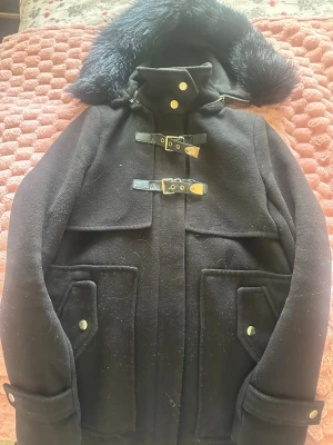 Svart duffelkappa med fuskpäls - Stilren svart duffelkappa med stora fickor, dekorativa spännen i metall och läderdetaljer. Kappan har en huva med mjuk fuskpäls och hög krage med tryckknappar. Perfekt för kalla dagar och ger en cool look.