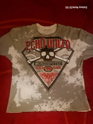 Grå och vit Ecko Unltd t-shirt - Cool t-shirt från Ecko Unltd i grått och vitt med stort tryck framtill. T-shirten har korta ärmar och ett unikt, splash-liknande mönster. Materialet är mjuk bomull och passformen är normal. Perfekt för dig som gillar streetwear och statement-plagg.