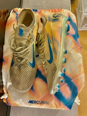 Nike Mercurial vapor 16 elite KM, AG - Skick 7/10! Säljer ett par Nike Mercurial fotbollsskor i beige med blå detaljer och sula. Skorna har en strömlinjeformad design, snörning och mönstrad ovandel i syntetmaterial. Perfekta för dig som vill ha grym touch och kontroll på planen.