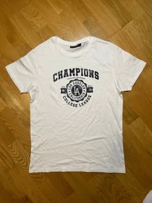 Vit t-shirt med tryck Reaction - Säljer denna vita t-shirt med svart tryck på bröstet där det står 'Champions' och 'Los Angeles California College League' i storlek . Har inprincip aldrig använt den (har endast provat den en gång). Tröjan har en klassisk rund hals och korta ärmar. Perfekt för en avslappnad och sportig stil. Vid frågor, extra bilder eller liknande är det bara att kontakta mig!