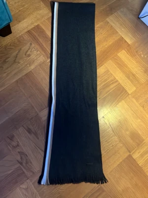 Svart halsduk med vit rand från BOSS - Snygg svart halsduk från BOSS med en vit och beige rand längs ena sidan. Halsduken har fransar i ändarna och är stickad i ett mjukt material. Perfekt accessoar för att lyfta din outfit.