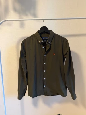 Mörkgrön skjorta från Ralph Lauren - Klassisk mörkgrön skjorta från Ralph Lauren med orange broderad logga på bröstet. Skjortan har långa ärmar, knappar framtill och button-down krage. Perfekt för en stilren och avslappnad look.