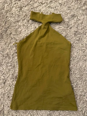 Olivgrön halterneck tubtopp - Säljer en olivgrön tubtopp med unik halterneck och hög krage. Toppen är ärmlös och har en tight passform som framhäver figuren. Perfekt för dig som vill sticka ut med en cool och trendig look.