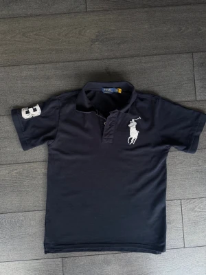 Mörkblå piké från Polo Ralph Lauren - Klassisk mörkblå pikétröja från Polo Ralph Lauren med vit broderad logga på bröstet och siffran 3 på ärmen. Skjortan har korta ärmar, krage och knappar vid halsen. Perfekt för en stilren och sportig look.