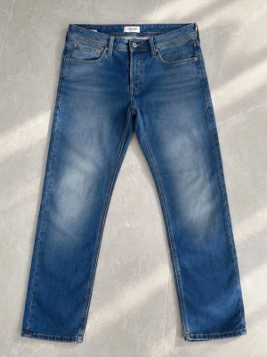 Blå slim fit jeans med ljus tvätt - Snygga blå jeans med slim fit  passform och ljus tvätt på låren. Klassisk femficksmodell med knappgylf och kontrastsömmar. Jeansen har en patch bak i midjan och diskret broderi på bakfickorna. Perfekta till vardagslooken.