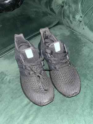 Ultraboost size 39 - Säljer ett par helsvarta sneakers med stickad ovandel och matchande svarta snören. Skorna har en sportig look med plastdetaljer på sidorna och en bekväm, dämpad sula. Perfekta för dig som gillar stilrena och bekväma skor.