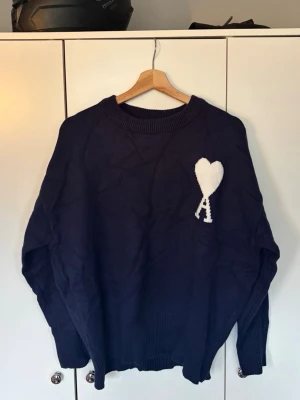 Mörkblå stickad tröja med hjärtdetalj - Mysig mörkblå stickad tröja med rund halsringning och ribbade muddar. På bröstet finns en vit stickad detalj i form av ett hjärta med bokstaven A under. Perfekt för dig som gillar enkel men snygg design. Storlek S. Helt ny 