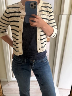Randig kort kofta i offwhite och svart - Säljer en söt, stickad kofta i offwhite med svarta horisontella ränder. Koftan har rund hals, knappar framtill och trekvartsärmar. Storlek 38 men sitter som 36.