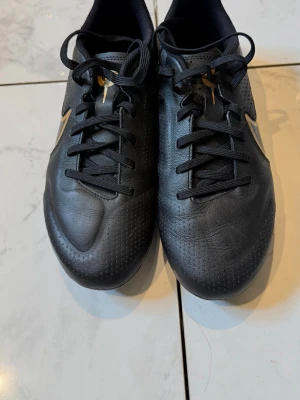 Svarta fotbollsskor med guldiga detaljer - Snygga svarta fotbollsskor med guldiga detaljer och klassisk snörning. Skorna har en låg profil och är tillverkade i syntetmaterial med texturerad yta för bättre bollkontroll. Perfekta för dig som vill ha stilrena och funktionella skor på planen.