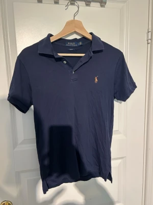Marinblå piké från Polo Ralph Lauren - Klassisk marinblå pikétröja från Polo Ralph Lauren i slim fit-modell. Tröjan har korta ärmar, krage och knappar framtill samt den ikoniska broderade loggan på bröstet. Tillverkad i mjuk bomull för en skön känsla.