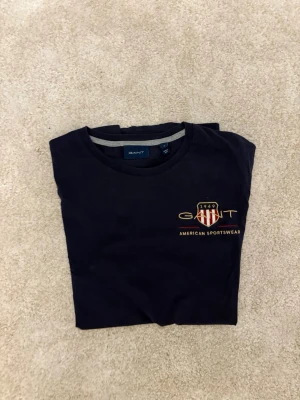 Mörkblå GANT t-shirt med tryck - Snygg mörkblå t-shirt från GANT med klassiskt tryck och logga på bröstet. T-shirten har rund hals och korta ärmar, perfekt för en clean och sportig look. Tillverkad i mjuk bomull för skön känsla hela dagen.