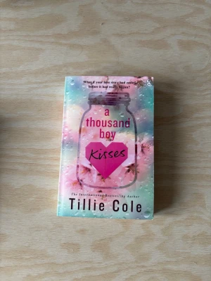 A Thousand Boy Kisses av Tillie Cole - En romantisk roman på engelska med färgstarkt omslag i pastelltoner och motiv av en glasburk och rosa hjärta. Perfekt för dig som gillar känslosamma berättelser om kärlek och ungdom. Mjukpärm och smidig att ta med sig.