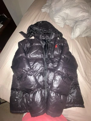 Svart pufferjacka från Polo Ralph Lauren - Snygg svart pufferjacka från Polo Ralph Lauren med röd logga på bröstet. Jackan har huva, dragkedja framtill och stora vadderade sektioner som håller dig varm. Materialet är glansigt och ger en modern look. Perfekt för kalla vinterdagar.