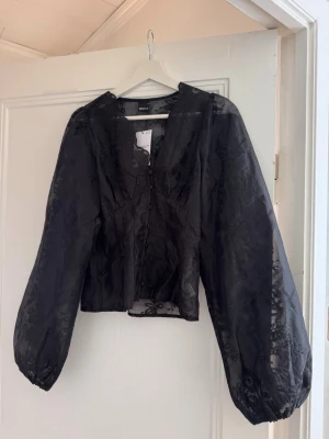 Gina blus - Trendig svart blus i transparent material med puffiga långärmar och knappar framtill. Blusen har en lätt glansig finish och subtilt blommönster i tyget. Perfekt för att lyfta en outfit med en edgy touch. Oanvänd med prislapp