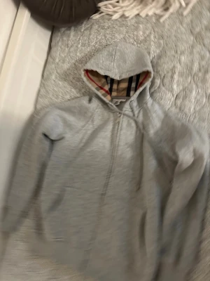 Grå hoodie med dragkedja och huva - Säljer en grå hoodie med dragkedja och huva. Insidan av huvan har ett klassiskt rutigt mönster i beige, rött och svart. Hoodien har broderad logga på bröstet och är tillverkad i mjukt sweatshirtmaterial. Perfekt för en chill och stilren look.
