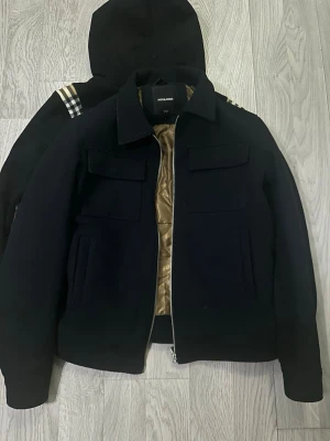 Jack & Jones kappa - Stilren svart jacka från Jack & Jones med två stora bröstfickor och dragkedja framtill. Jackan har en snygg guldig insida och klassisk krage. Perfekt för dig som vill ha en enkel men trendig look.