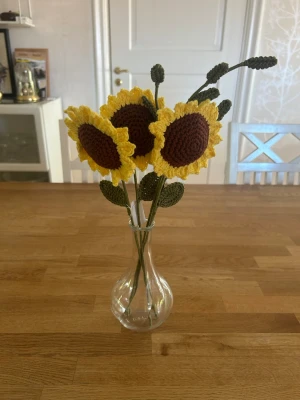 Virkat solrosbukett i glasvas - Unika handgjorda virkade solrosor i brunt och gult garn med gröna stjälkar och blad. Blommorna står i en enkel, genomskinlig glasvas. Perfekt som dekoration till hemmet eller som present till någon speciell.