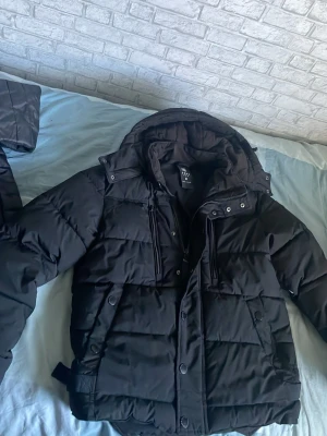 Svart quiltad pufferjacka med huva - Säljer en svart pufferjacka med huva och quiltad design. Jackan har långa ärmar, dragkedja och knappar framtill samt praktiska fickor med dragkedja. Perfekt för kalla dagar och riktigt skön att bära. Storlek M men skulle säga mer S