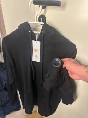 Svart hoodie från C.P. Company - Säljer en svart hoodie från C.P. Company med den ikoniska linsen på ärmen. Hoodien har snörning i huvan och en klassisk, avslappnad passform. Perfekt för dig som gillar streetwear och vill ha något stilrent och bekvämt.