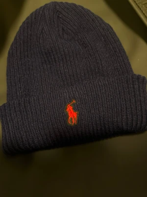 Mörkblå  mössa Polo - Säljer en mörkblå ribbstickad mössa från Polo Ralph Lauren med klassisk röd broderad logga framtill. Mössan har uppvikt kant och är tillverkad i mjukt, värmande material som passar perfekt för kyliga dagar.