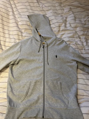 Grå hoodie från Polo Ralph Lauren - Snygg ljusgrå hoodie från Polo Ralph Lauren med dragkedja, två fickor fram och justerbar huva med snören. Klassisk logga broderad på bröstet. Perfekt för en chill och stilren look. Tillverkad i mjukt bomullsmaterial.