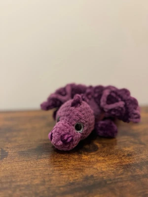 🐉 Handvirkad liten drake 💜 - Söt handvirkad drake i mjukt, lila garn med stora svarta ögon och detaljerade vingar. Draken har en rund kropp och små horn på huvudet. Perfekt som dekoration eller gosedjur för den som gillar fantasy och unika prylar.