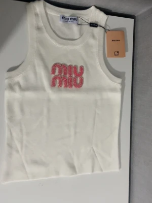  Miu Miu linnetopp - Supersnygg vit stickad linnetopp från Miu Miu med rosa pärlbroderat logotyp framtill. Toppen är ärmlös och har en rund halsringning. Perfekt för dig som vill ha en trendig och exklusiv look med en touch av lyx.