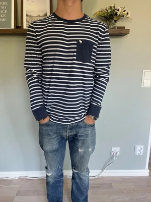 Abercrombie & Fitch Randig Långärmad Tröja Navy/White S - Abercrombie & Fitch långärmad tröja i marinblått och vitt med klassiskt randigt mönster. Stilren modell med bröstficka och den ikoniska älg-loggan. Tidlös design som passar perfekt till jeans, chinos eller shorts.  Skick: fint skick Storlek: S Färg: Navy/White Passform: regular fit  
