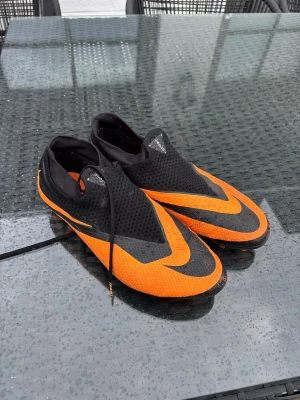 Svart-orangea Nike fotbollsskor - Säljer ett par grymma fotbollsskor från Nike i svart och orange. Skorna har en modern, strömlinjeformad design med stora Swoosh-loggor och snörning på sidan. Ovandelen är i mesh-liknande material för bra ventilation och passform.