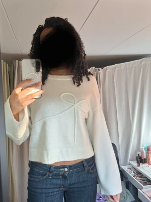 Vit cropped stickad tröja med rosett - Säljer en vit, cropped stickad tröja med en dekorativ rosett framtill. Tröjan har långa ärmar, rund halsringning och ribbade muddar vid ärmslut och nederkant. Perfekt för dig som gillar en söt och enkel stil.
