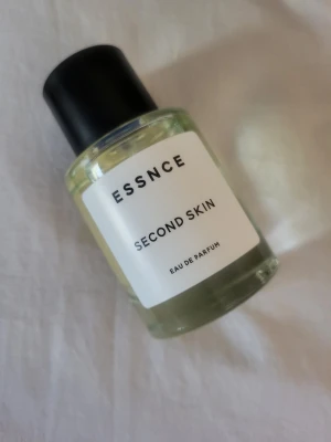 ESSNCE Second Skin Eau de Parfum - Fräsch parfym från ESSNCE med namnet Second Skin. Flaskan är rund och genomskinlig med svart lock och vit etikett. Innehållet är ljusgult och flaskan har en modern, minimalistisk design. Perfekt för dig som gillar stilrena dofter.