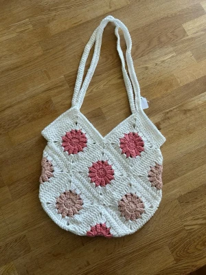 Virkad shoppingväska med blommor - Unik shoppingväska i virkat material med vita, rosa och beige toner. Väskan har dubbla axelband och ett mönster av virkade blommor i olika nyanser. Perfekt för dig som gillar boho-stil och vill ha en personlig accessoar.
