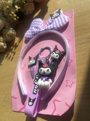 Lila håraccessoarset med Kuromi - Sött set med lila hårband, hårspännen och nyckelring, dekorerade med Kuromi-figurer och rutigt rosett. Perfekt för dig som gillar kawaii-stil och vill piffa upp frisyren med gulliga detaljer. Materialet är plast och tyg i lila och svart med vita och rosa inslag.