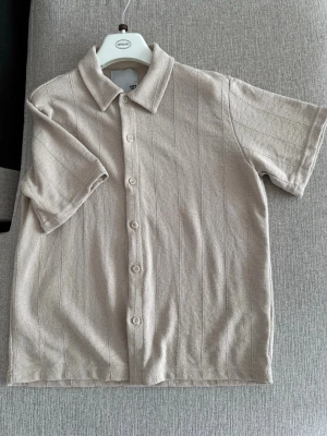 Beige stickad skjorta med knappar - Säljer en beige stickad skjorta med korta ärmar och klassisk krage. Skjortan har diskreta vertikala ränder och knäppning framtill. Perfekt för dig som gillar en avslappnad men ändå stilren look.