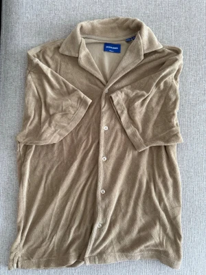 Beige frottéskjorta med kort ärm - Snygg beige skjorta i mjuk frotté med korta ärmar och klassisk krage. Skjortan har knäppning framtill med vita knappar och en avslappnad passform. Perfekt för en chill och trendig look.