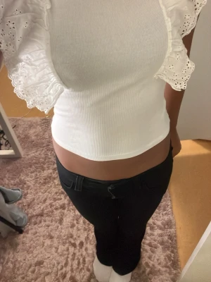 Vit croppad topp med volangärmar - Säljer en vit croppad topp med ribbad struktur och breda volangärmar i broderad spets. Toppen är ärmlös och har en tight passform som framhäver midjan. Perfekt för dig som vill ha en söt och trendig look.