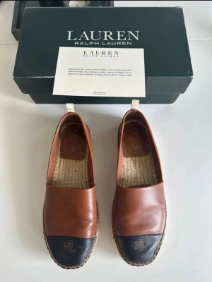 Espadrillor från Lauren Ralph Lauren modell Dorian - Snygga espadrillos i brunt läder med svart tåparti och RL-logga framtill. Skorna har en klassisk espadrillesula i flätad jute och en låg slip-on design. Perfekta för en avslappnad men stilren look. Små i storleken så passar storlek 39/40