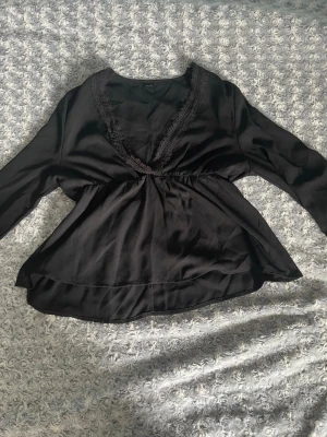 Svart blus med spetsdetalj - Snygg svart blus med djup v-ringning och spetskant längs urringningen. Blusen har lång ärm och markerad midja som ger en feminin siluett. Tillverkad i ett mjukt, lätt tyg som faller fint. Perfekt att styla med jeans eller kjol.