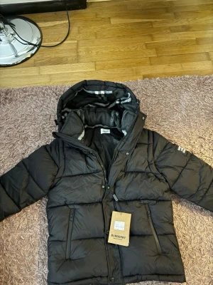 Svart pufferjacka från Burberry - Säljer en svart pufferjacka från Burberry med huva och dragkedja framtill. Jackan har två fickor med dragkedja och en patch på ärmen med texten 'BURBERRY LONDON ENGLAND'. Insidan av huvan har ett rutigt mönster. Perfekt för kalla vinterdagar.