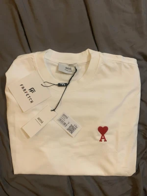 Vit t-shirt från Ami med rött hjärta - Säljer en stilren vit t-shirt från Ami med ett broderat rött hjärta och bokstaven A på bröstet. Klassisk passform och rund halsringning. Perfekt basplagg för dig som gillar enkel men snygg design.